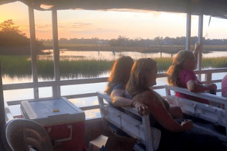 People seated on a boat at sunset, overlooking a calm river and distant trees.